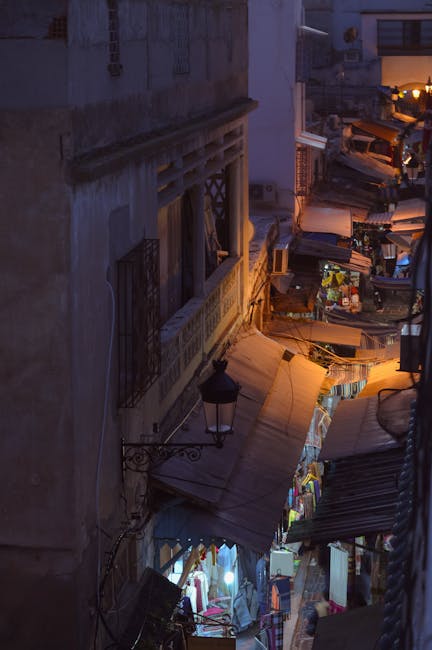 A densely packed urban outdoor scene at dusk, featuring narrow alleyways between tall, weathered buildings with textured facades. Small market stalls and makeshift awnings line the alleys, illuminated